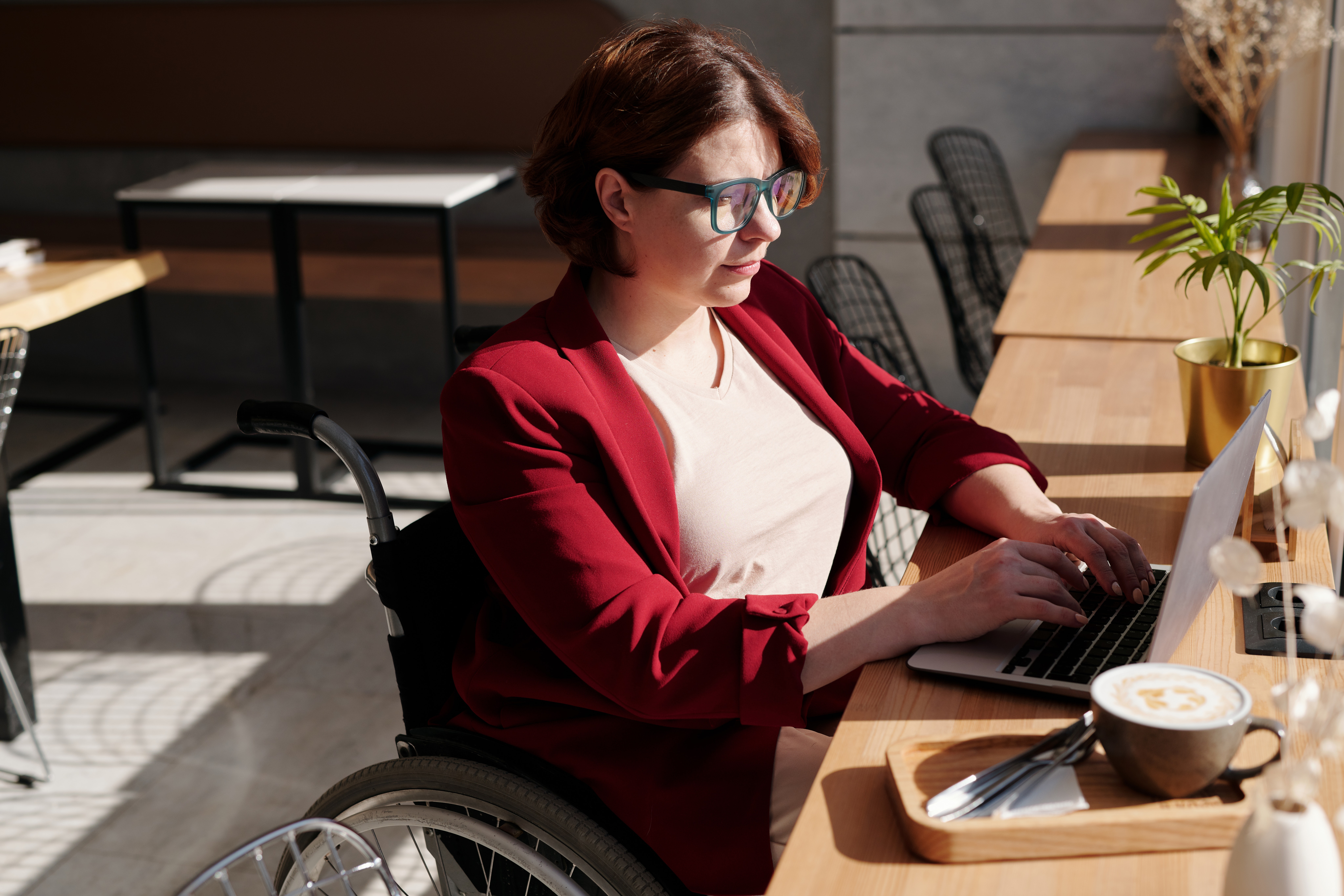 White woman in her mid-30's who is a wheelchair user sits in a café and works on her laptop while having a latte. She is wearing glasses with blue frames, a red blazer, and has red hair.